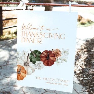 May include: A white Thanksgiving dinner sign with copper-colored text and a floral and pumpkin design. The sign reads "Welcome to our THANKSGIVING DINNER" and includes the family name and date.