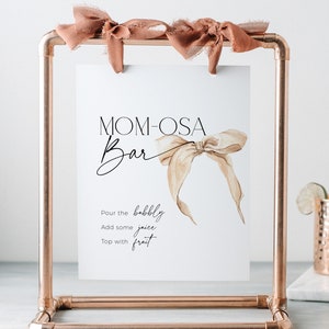 May include: A copper pipe stand with a white sign that says "MOM-OSA Bar" with a watercolour illustration of a brown ribbon bow. The sign also says "Pour the bubbly, Add some juice, Top with fruit".