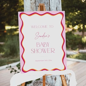 May include: Welcome to Sandra's Baby Shower sign with pink, orange, and white wavy borders. The sign is on an easel and says "Welcome to Sandra's Baby Shower" with the date "September 14th, 20xx".