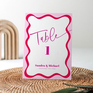 Könnte beinhalten: Rosa und rote Tischkartennummer mit wellenförmigem Rand. Die Karte zeigt "Tisch 1" und "Sandra & Michael 16. September 20xx".