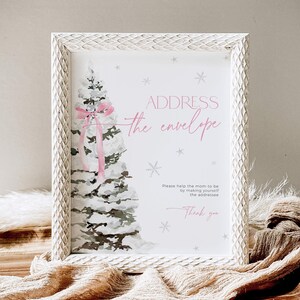 May include: A white frame with a white and gray watercolor illustration of a snow-covered Christmas tree with a pink bow. The text "ADDRESS the envelope" is written in pink and black. The text "Please help the mom to be by making yourself the addressee Thank you" is written in black.
