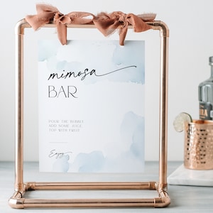 May include: A copper-colored metal frame holds a sign that reads "mimosa BAR" in black script. The sign has a watercolor blue background and instructions. A copper mug, a bottle, and a lime wedge are also visible.