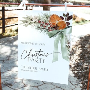 May include: A white sign with a watercolor illustration of a Christmas wreath with oranges, berries, and greenery. The text on the sign reads "Welcome to our Christmas Party, The Miller Family, December 18th, 20xx".