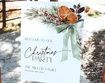 Könnte beinhalten: Ein weißes Schild mit einer Aquarell-Illustration eines Weihnachtskranzes mit Orangen, Beeren und Grün. Der Text auf dem Schild lautet "Willkommen zu unserer Weihnachtsfeier, Die Familie Miller, 18. Dezember 20xx".