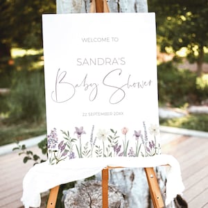 May include: White welcome sign with the text "Welcome to Sandra's Baby Shower" and the date "22 September 20XX". The sign is decorated with a floral border of purple and white flowers.