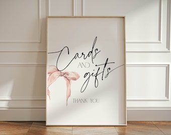 May include: A white sign with a gold frame and the text "Cards and gifts" in black cursive writing. A pink watercolor bow is to the left of the text. The text "Thank you" is below the bow.