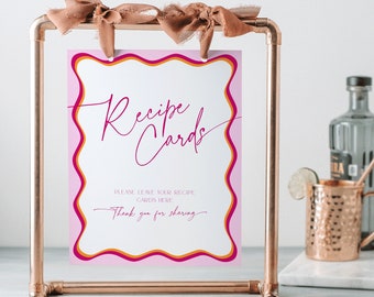 May include: Pink and orange recipe card sign with the text "Recipe Cards" in cursive script. The sign is on a copper pipe stand and says "Please leave your recipe cards here! Thank you for sharing."