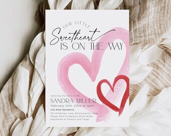 Op de afbeelding: Een babyshower-uitnodiging met een roze en rood aquarelhartontwerp. De tekst luidt "Onze kleine schat is onderweg" en bevat de naam Sandra Miller, de datum 14 februari 20xx om 16:00 uur, de locatie Lilly Rose Residence en het adres 301 Wildflower Lane, Bloomingville, Texas. RSVP naar Melanie 01234-56789. Geregistreerd bij Amazon en Target.