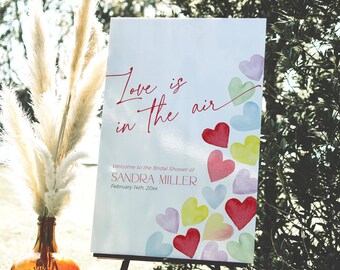 Puede incluir: Un letrero blanco con el texto "Love is in the air" en escritura roja, y un mensaje de bienvenida para la despedida de soltera de Sandra Miller. Corazones de acuarela coloridos decoran el letrero. El letrero se muestra en un caballete negro.