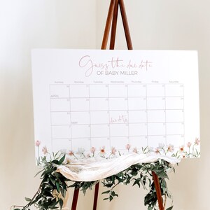 May include: A white calendar with a floral border, featuring a pink script that says "Guess the due date of Baby Miller". The calendar is set up for April and May, with the due date marked on the calendar.