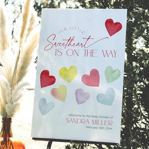 May include: A white sign with red and pink text that reads "Our Little Sweetheart Is On The Way". The sign also includes a watercolor illustration of hearts in various colors. The sign is on an easel and is in front of a vase with dried pampas grass.