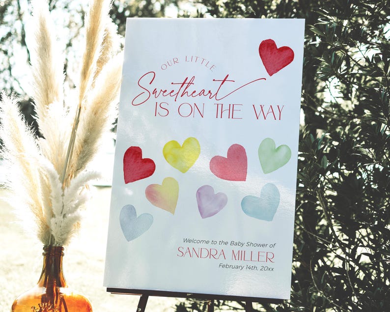 May include: A white sign with red and pink text that reads "Our Little Sweetheart Is On The Way". The sign also includes a watercolor illustration of hearts in various colors. The sign is on an easel and is in front of a vase with dried pampas grass.