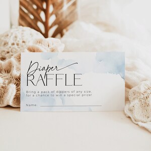 May include: A white rectangular card with the words "Diaper RAFFLE" in black script and sans-serif font. The card has a watercolor blue wash design. Text below reads "Bring a pack of diapers of any size... for a chance to win a special prize!"