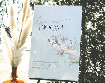 May include: A white sign with pink floral watercolour artwork and the text "love in BLOOM" in pink and black lettering. The sign also includes the text "Welcome to the Bridal Shower of SANDRA MILLER September 14th, 20xx".