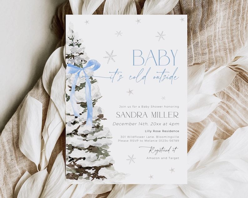 May include: A blue and white baby shower invitation with a watercolor illustration of a snow-covered pine tree with a blue ribbon. The text reads "Baby it's cold outside" and includes details for the event, including the date, time, location, and RSVP information. The invitation also includes the text "Registered at Amazon and Target".