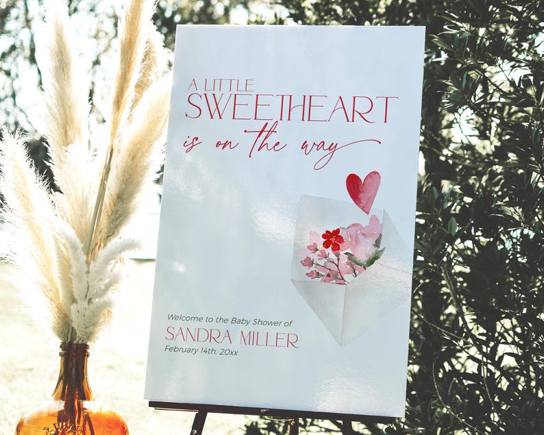 May include: A white sign with red text announcing a baby shower for Sandra Miller. The text reads "A little sweetheart is on the way". A watercolor illustration of a pink envelope with a red heart and pink flowers is in the bottom right corner.