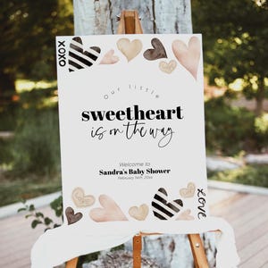 K&ouml;nnte beinhalten: Ein wei&szlig;es Schild mit schwarzem Text, der "Unser kleines Herzchen ist unterwegs." lautet. Willkommen zur Babyparty von Sandra, 4. Februar 20xx. Das Schild ist mit schwarzen und wei&szlig;en Herzen sowie rosa und braunen Herzen verziert.