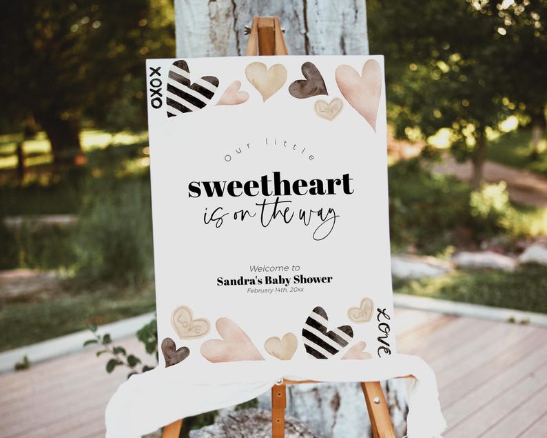 K&ouml;nnte beinhalten: Ein wei&szlig;es Schild mit schwarzem Text, der "Unser kleines Herzchen ist unterwegs." lautet. Willkommen zur Babyparty von Sandra, 4. Februar 20xx. Das Schild ist mit schwarzen und wei&szlig;en Herzen sowie rosa und braunen Herzen verziert.