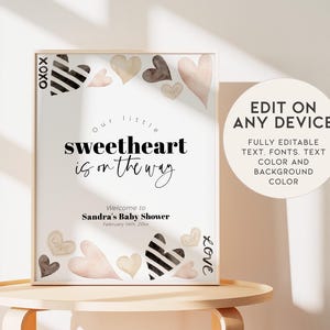 K&ouml;nnte beinhalten: Druckbarer Babyparty-Schild in den Farben Rosa, Wei&szlig; und Schwarz. Das Schild zeigt ein Herzdesign und den Text "Unser kleiner Liebling ist unterwegs".