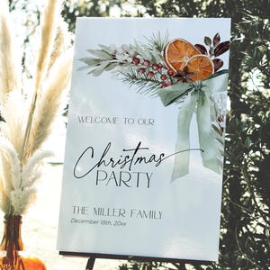 May include: A white sign with a watercolor illustration of a green wreath with orange slices, berries, and a green bow. The text on the sign reads "Welcome to our Christmas Party The Miller Family December 18th, 20xx".