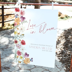 Op de afbeelding: Een wit bord met roze bloemmotieven en vlinders. Het bord zegt "Love in Bloom" en "Welkom op de bruidsdouche van Sandra Miller, 14 juni 20xx".