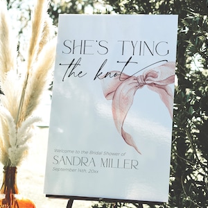 Può includere: Un cartello bianco con la scritta "SHE'S TYING the knot" e un'illustrazione di fiocco ad acquerello. Sotto, si legge "Welcome to the Bridal Shower of SANDRA MILLER September 14th, 20xx". Il cartello è su un cavalletto nero.