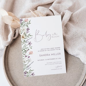 May include: A white baby shower invitation with a floral border. The text reads "Baby in Bloom" and includes the name "Sandra Miller" and details for the event.