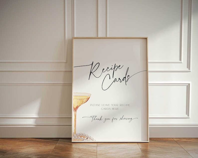 May include: A white sign with a gold frame, featuring a black calligraphy script that reads "Recipe Cards". Below the text is a watercolor illustration of a champagne flute with a pearl necklace draped around the stem. The sign also includes the text "Please leave your recipe cards here. Thank you for sharing."