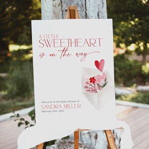 May include: A white sign with red text announcing a baby shower for Sandra Miller. The sign reads "A little sweetheart is on the way." and includes a watercolor illustration of a pink envelope with a red heart and flowers.