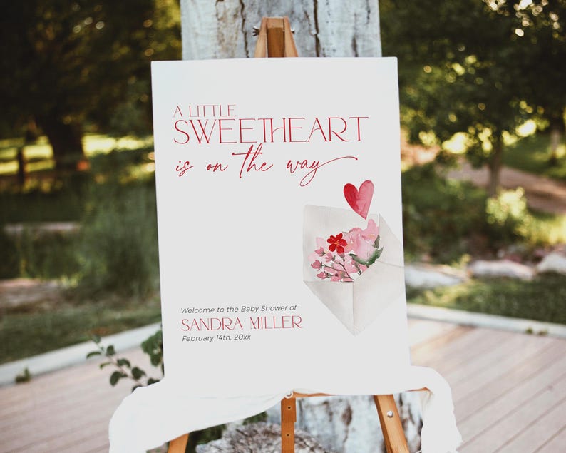 May include: A white sign with red text announcing a baby shower for Sandra Miller. The sign reads "A little sweetheart is on the way." and includes a watercolor illustration of a pink envelope with a red heart and flowers.