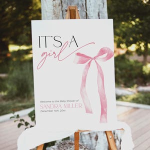 May include: It's a girl baby shower sign featuring a pink watercolour bow and the text "Welcome to the Baby Shower of Sandra Miller December 14th 20xx".