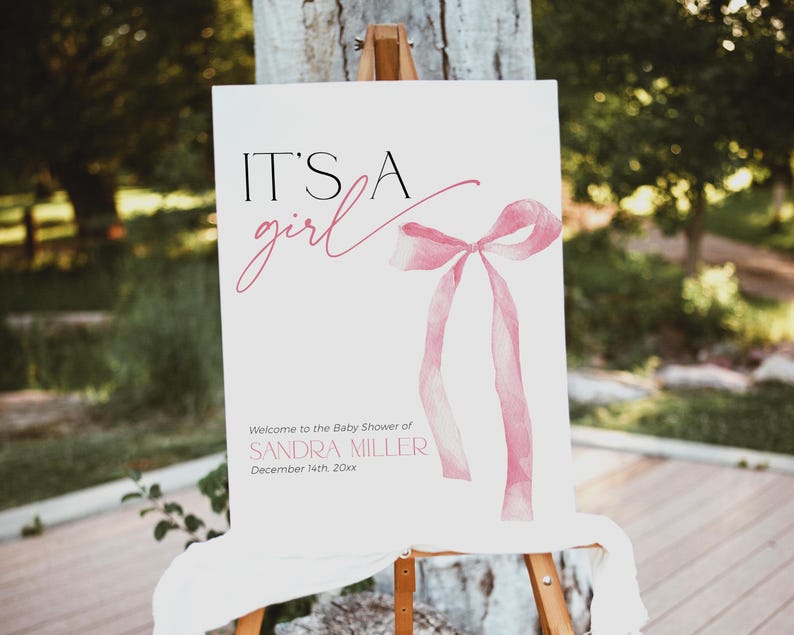 May include: It's a girl baby shower sign featuring a pink watercolour bow and the text "Welcome to the Baby Shower of Sandra Miller December 14th 20xx".