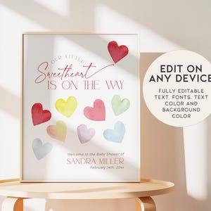 May include: A printable baby shower sign with the text "Our Little Sweetheart Is On The Way" in pink script font. The sign features a pink heart with a tail and a rainbow of watercolor hearts.