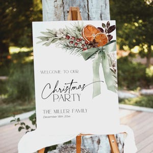 May include: A white sign with a watercolor illustration of a green wreath with oranges, berries, and a green bow. The text on the sign reads "Welcome to our Christmas Party The Miller Family December 18th, 20xx".