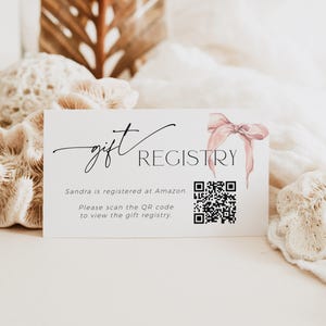 May include: A white card with the text "gift REGISTRY" in black script font. The text "Sandra is registered at Amazon. Please scan the QR code to view the gift registry." is printed below the title. A pink watercolor bow is in the upper right corner of the card. A black and white QR code is in the center of the card.