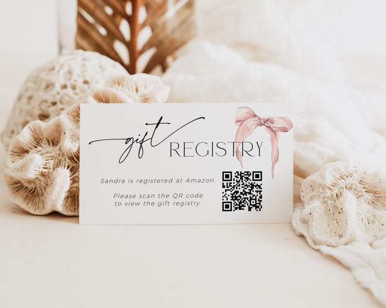 May include: A white card with the text "gift REGISTRY" in black script font. The text "Sandra is registered at Amazon. Please scan the QR code to view the gift registry." is printed below the title. A pink watercolor bow is in the upper right corner of the card. A black and white QR code is in the center of the card.