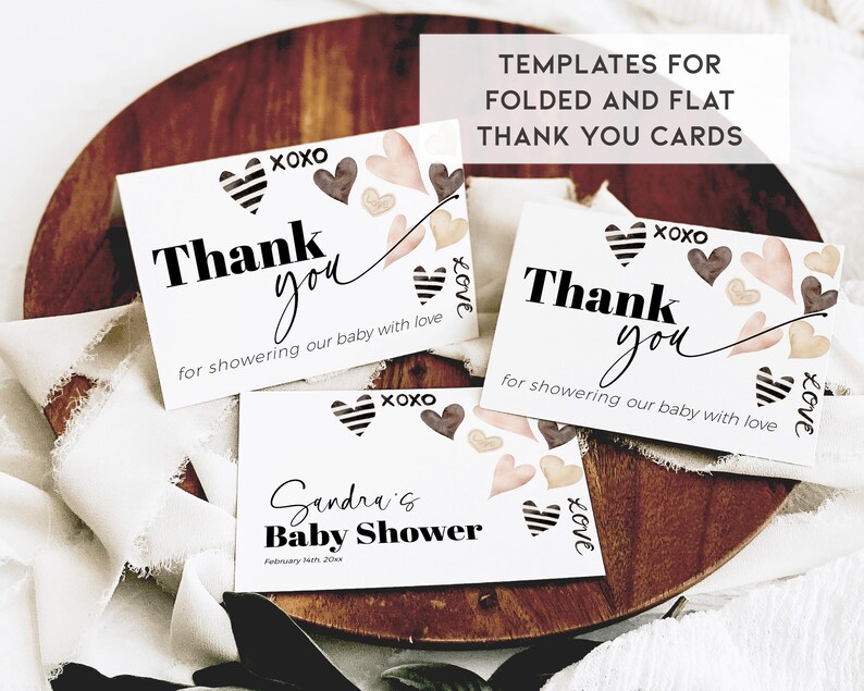 May include: Two white thank you cards with black text and brown and pink watercolour hearts. The text on the cards reads "Thank you for showering our baby with love." The cards are on a wooden surface with a white fabric background. The text "TEMPLATES FOR FOLDED AND FLAT THANK YOU CARDS" is at the top of the image.