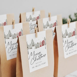 May include: Kraft paper gift bags with attached tags. The tags feature a winter scene with houses and trees, and the words "Merry Christmas" in elegant script. Each tag is secured with a small wooden clothespin. The text "with love" and "THE MILLER FAMILY" are also visible.