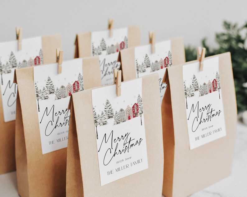 May include: Kraft paper gift bags with attached tags. The tags feature a winter scene with houses and trees, and the words "Merry Christmas" in elegant script. Each tag is secured with a small wooden clothespin. The text "with love" and "THE MILLER FAMILY" are also visible.