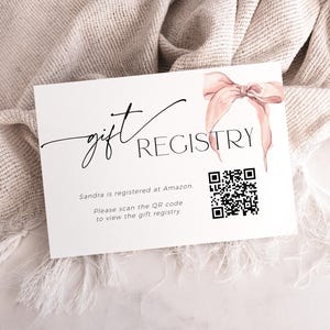May include: A white card with black text that reads "gift registry". A pink ribbon is in the top right corner. A black and white QR code is below the text. The text below the QR code reads "Sandra is registered at Amazon. Please scan the QR code to view the gift registry."
