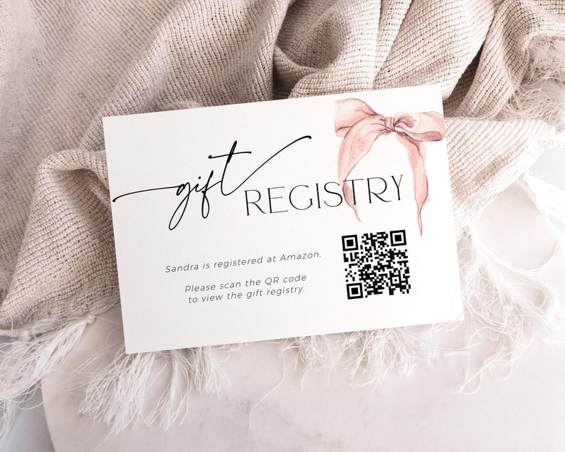 May include: A white card with black text that reads "gift registry". A pink ribbon is in the top right corner. A black and white QR code is below the text. The text below the QR code reads "Sandra is registered at Amazon. Please scan the QR code to view the gift registry."