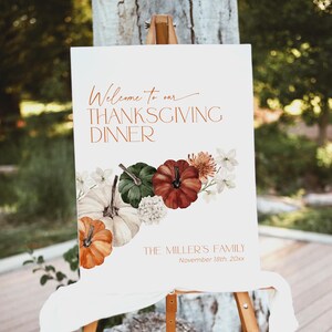 May include: A white Thanksgiving dinner sign on an easel. The sign has the text "Welcome to our THANKSGIVING DINNER" in copper-colored script. It features watercolor pumpkins in orange, green, and red, with white flowers. The sign also includes the family name and date.