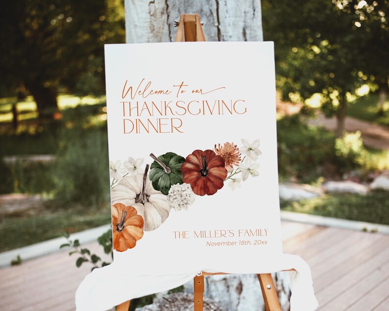 May include: A white Thanksgiving dinner sign on an easel. The sign has the text "Welcome to our THANKSGIVING DINNER" in copper-colored script. It features watercolor pumpkins in orange, green, and red, with white flowers. The sign also includes the family name and date.