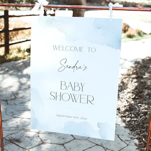 May include: A white sign with blue watercolor accents announcing "Welcome to Sandra's Baby Shower" with the date "September 14th, 20xx". The sign is hanging from a copper pipe stand.