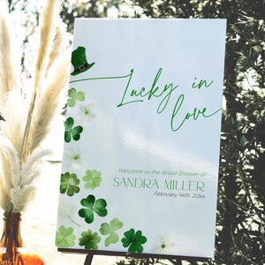 May include: A white sign with green shamrocks and a green leprechaun hat. The sign says "Lucky in Love" and "Welcome to the Bridal Shower of Sandra Miller, February 14th, 20xx".