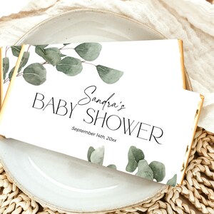 May include: White card with gold trim featuring a watercolor illustration of eucalyptus leaves. The card reads "Sandra's Baby Shower" and "September 14th, 20xx".
