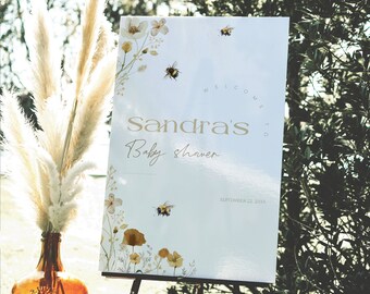 May include: A white baby shower welcome sign with floral and bee illustrations. The sign reads "Sandra's Baby shower" in gold lettering, with the date "September 22, 20XX" at the bottom. The sign is displayed on a black easel.