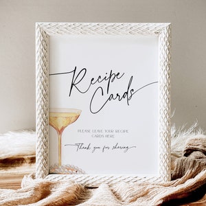May include: White framed sign with the text "Recipe Cards" in black cursive script. A glass of champagne with a pearl necklace is pictured below the text. The sign reads "Please leave your recipe cards here. Thank you for sharing."