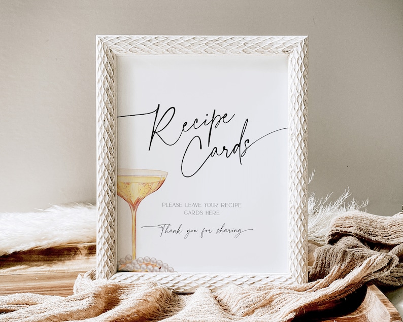 May include: White framed sign with the text "Recipe Cards" in black cursive script. A glass of champagne with a pearl necklace is pictured below the text. The sign reads "Please leave your recipe cards here. Thank you for sharing."