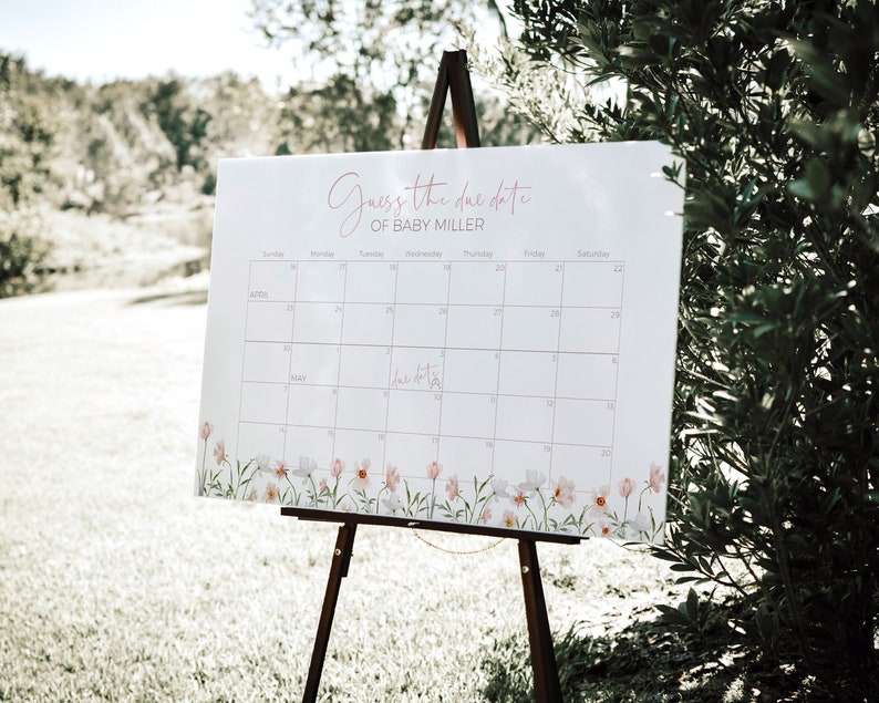 May include: A white calendar with a floral border, set up on an easel. The calendar is titled "Guess the due date of Baby Miller" and has the months of April and May displayed.